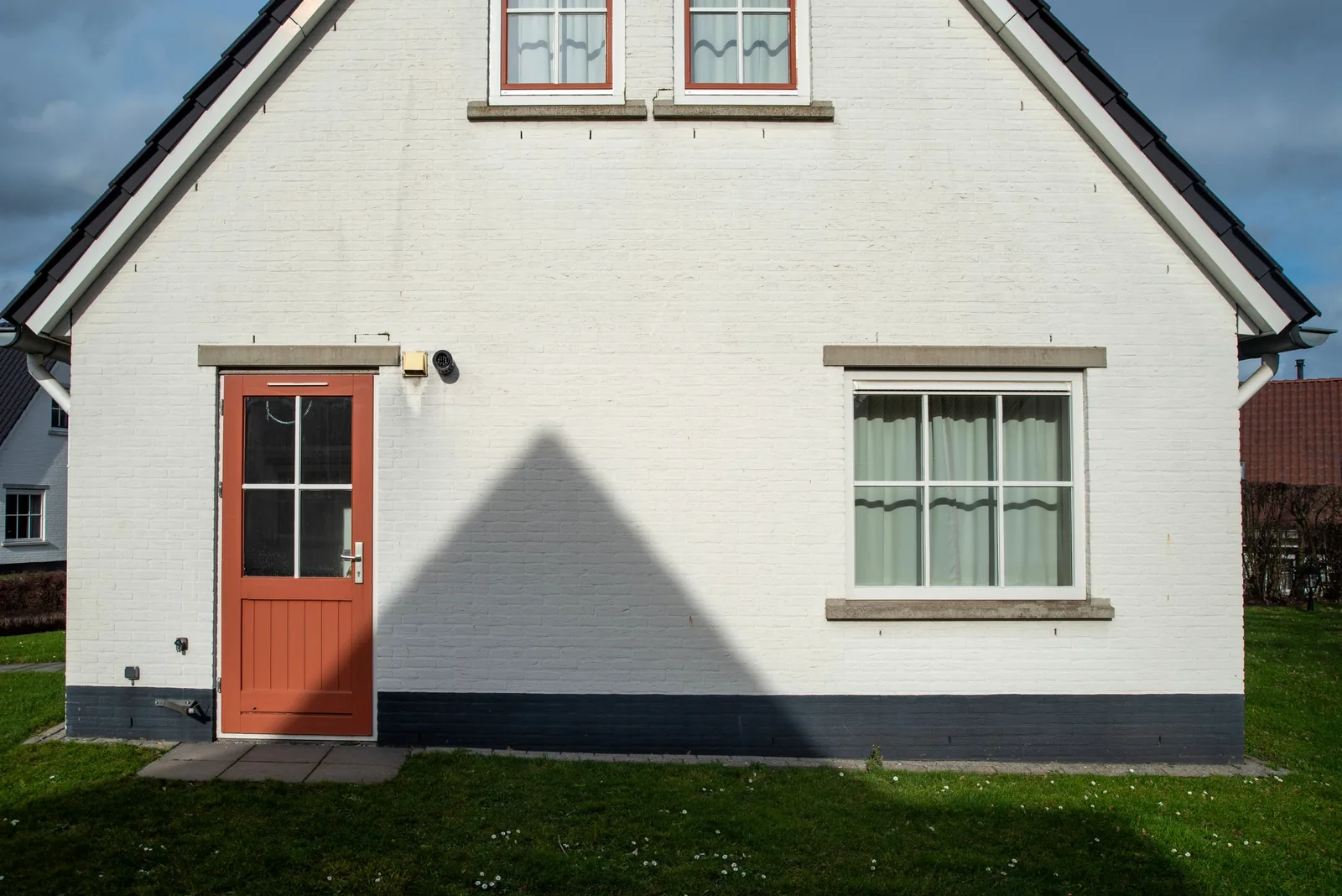 White gabled house with a red door and its shadow on the lawn